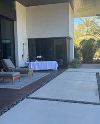 Relaxing modern patio with cushioned wooden lounge chairs, outdoor shower and white-covered massage table on a wooden deck, large concrete pavers edged with river rocks and palm trees beyond.