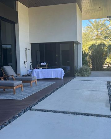 Relaxing modern patio with cushioned wooden lounge chairs, outdoor shower and white-covered massage table on a wooden deck, large concrete pavers edged with river rocks and palm trees beyond.