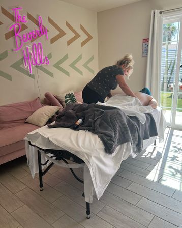 Therapist giving a massage on a portable table in a bright Beverly Hills–style room with a pink neon wall sign, blush sofa, textured gray blanket, and sliding glass door showing palm trees