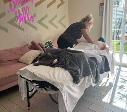 Therapist giving a massage on a portable table in a bright Beverly Hills–style room with a pink neon wall sign, blush sofa, textured gray blanket, and sliding glass door showing palm trees