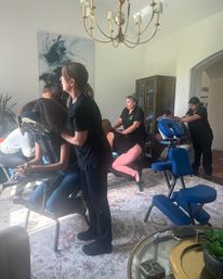 Group on-site chair massage in a bright residential living room — therapists giving portable chair massages on blue chairs under a brass chandelier with abstract wall art visible.
