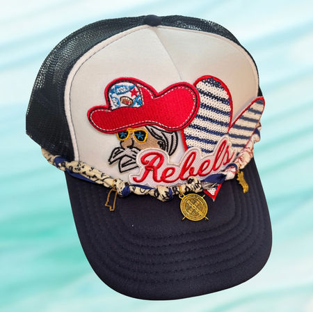 Embroidered white-and-navy trucker hat with a red cowboy patch, 'Rebels' script, striped heart, braided bandana cord and gold charms on an aqua background.