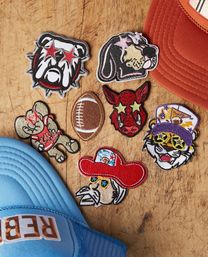 Colorful embroidered mascot and sports patches—star-eyed bulldog, sleeping hound, elephant, stitched football, red bull face, cowboy with red hat, and tiger in a purple cap—arranged on a scratched wooden table beside blue and orange trucker hats.