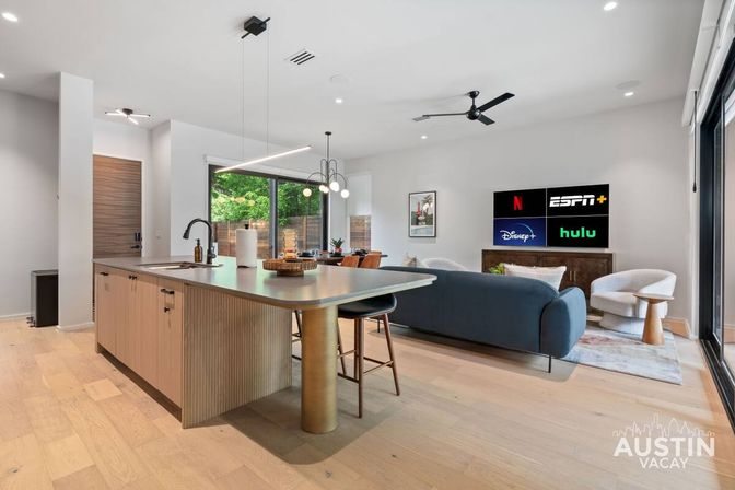 Bright modern open-plan kitchen and living room in an Austin vacation rental — wood island with brass leg, navy sofa, sliding glass doors, and TV displaying streaming apps