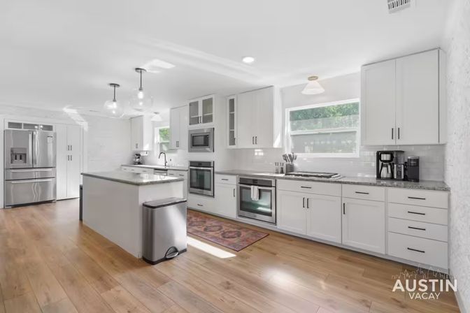 Bright modern white vacation-rental kitchen with island, granite counters, pendant lights, stainless-steel fridge and ovens, hardwood floors and coffee station