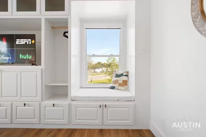 Sunny modern interior with white built-in cabinetry and media wall, cozy window seat with cushion overlooking a tree-lined street and hardwood floors