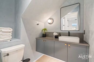 Cozy modern vacation-rental bathroom with gray floating vanity and gold knobs, white rectangular vessel sink with black faucet, round wall sconce, stacked white towels on a shelf, and a compact toilet under an angled ceiling.