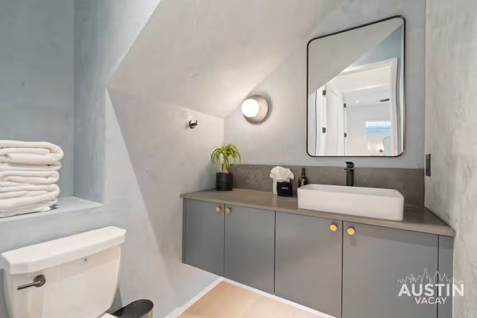 Cozy modern vacation-rental bathroom with gray floating vanity and gold knobs, white rectangular vessel sink with black faucet, round wall sconce, stacked white towels on a shelf, and a compact toilet under an angled ceiling.