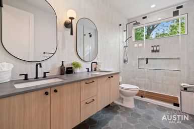 Sleek contemporary bathroom with double sinks and oval mirrors, black matte faucets on a gray countertop over wood-tone cabinets, hexagon tile floor, and a glass walk-in shower with rainfall showerhead and a tree-lined window.