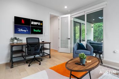 Bright modern Austin living room-office with wall-mounted TV displaying Netflix, ESPN+, Disney+ and Hulu logos, industrial desk and rolling chair, blue armchair by sliding glass doors to a green balcony, wooden coffee table and orange accent rug.