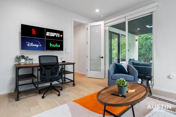 Bright modern Austin living room-office with wall-mounted TV displaying Netflix, ESPN+, Disney+ and Hulu logos, industrial desk and rolling chair, blue armchair by sliding glass doors to a green balcony, wooden coffee table and orange accent rug.