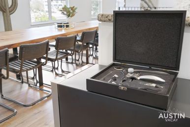 Open black wine accessory kit with corkscrew, stopper and pourer on a kitchen island overlooking a bright modern dining area with a long wooden table and metal‑frame chairs