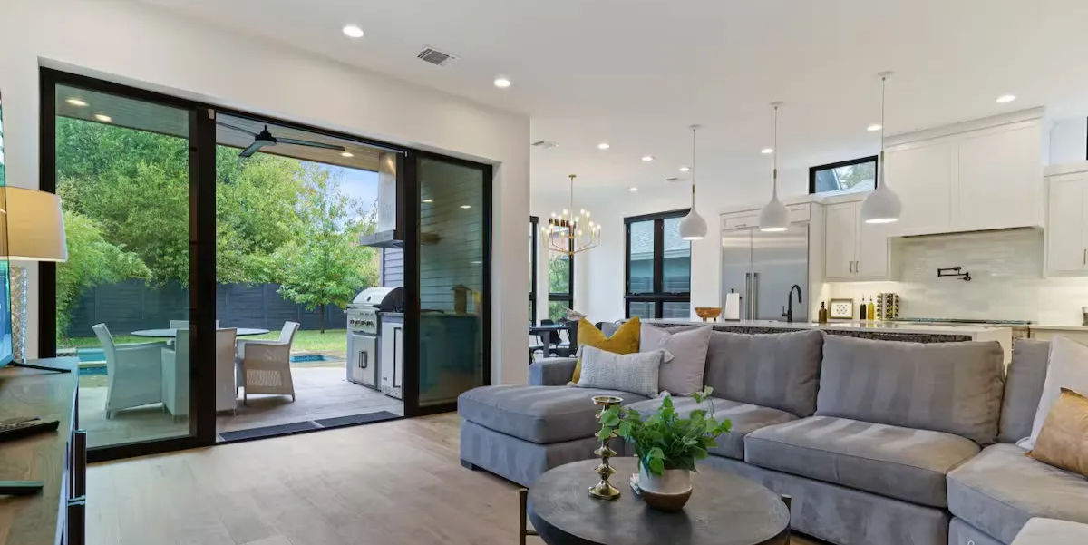 Sunlit modern open-plan living room and kitchen with a gray sectional, round coffee table and plants, pendant lights and hardwood floors, large sliding glass doors opening to a leafy backyard patio with grill and outdoor seating — ideal for indoor-outdoor entertaining.