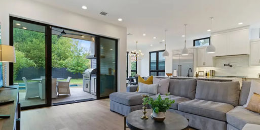 Sunlit modern open-plan living room and kitchen with a gray sectional, round coffee table and plants, pendant lights and hardwood floors, large sliding glass doors opening to a leafy backyard patio with grill and outdoor seating — ideal for indoor-outdoor entertaining.