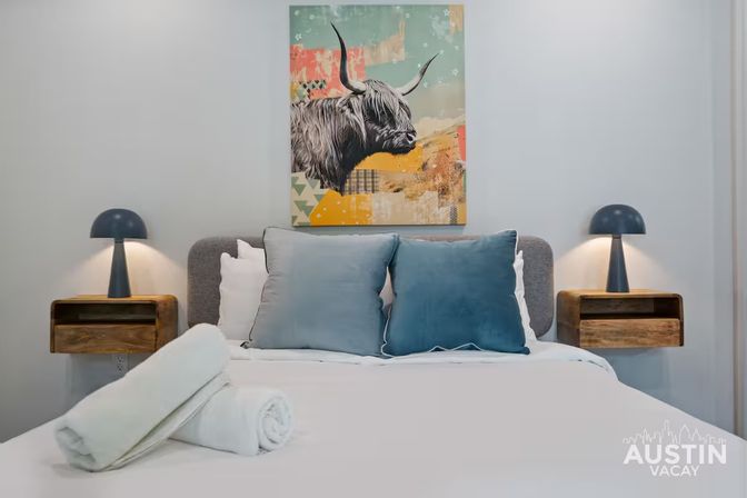Modern Austin vacation rental bedroom with white linens, two blue throw pillows, rolled white towels, wooden floating nightstands with navy dome lamps, and colorful highland cow wall art.