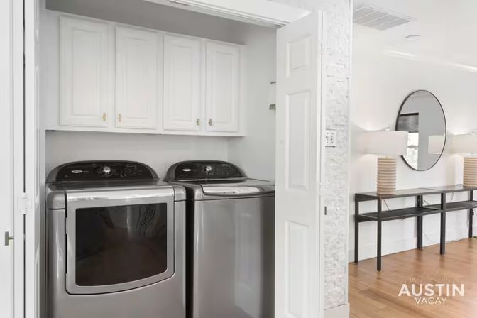 Modern laundry nook with stainless steel washer and dryer in a white closet, next to hardwood entryway with round mirror and console table.