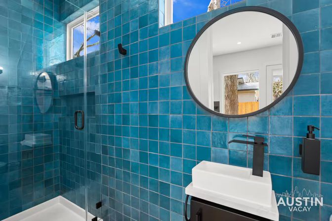 Vibrant teal-tiled bathroom in a modern vacation rental, with a glass shower, round mirror reflecting a window and tree, white sink and matte-black faucet and soap dispenser.