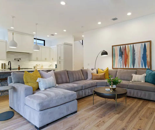 Modern open-concept living room and kitchen with a plush gray L-shaped sectional, mustard and teal accent pillows, round brass coffee table, hardwood floors, white shaker cabinets, pendant lights and colorful abstract wall art.