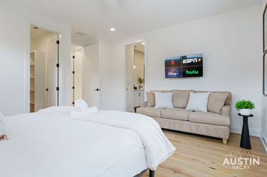 Bright modern vacation rental bedroom with white bed and rolled towel, beige sofa with pillows, wall-mounted TV showing streaming app logos, hardwood floors and open doorway to bathroom/closet.