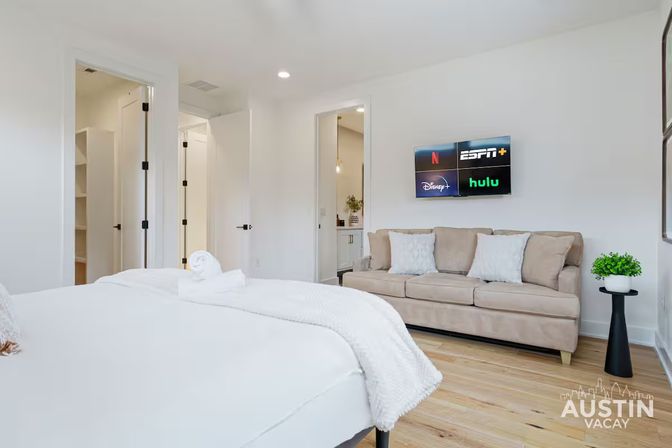 Bright modern vacation rental bedroom with white bed and rolled towel, beige sofa with pillows, wall-mounted TV showing streaming app logos, hardwood floors and open doorway to bathroom/closet.