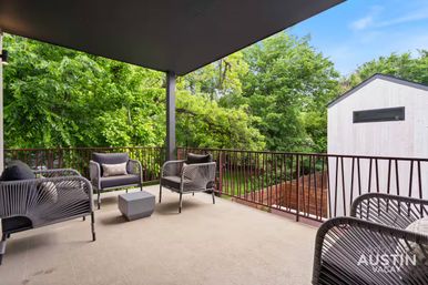 Relaxing modern covered balcony with woven lounge chairs and a cube table overlooking lush green trees and a neighboring light‑colored home.