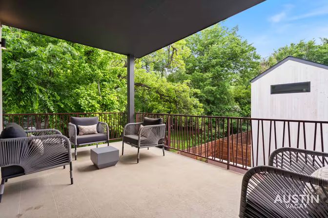 Relaxing modern covered balcony with woven lounge chairs and a cube table overlooking lush green trees and a neighboring light‑colored home.