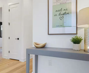 Modern entryway with gray console table on light oak floors, wooden bowl, small potted plant and lamp, framed “meet me in Austin” print on a white wall.