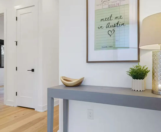 Modern entryway with gray console table on light oak floors, wooden bowl, small potted plant and lamp, framed “meet me in Austin” print on a white wall.