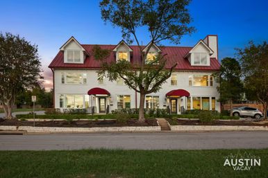 Charming three-story white guesthouse in Austin at dusk with a red metal roof, red awnings, lit windows, central oak tree and street-front landscaping