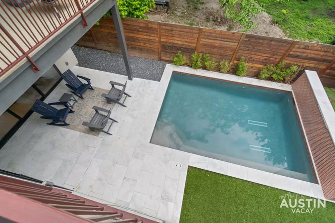 Aerial view from a balcony of a modern backyard with a rectangular plunge pool, light stone patio, four Adirondack chairs on a patterned rug, wooden privacy fence and green turf.
