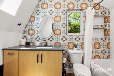 Bright modern bathroom with sloped ceiling and colorful geometric tile accent wall (orange, gray, blue starburst pattern), light wood vanity with black countertop and round mirror, small window showing green trees, white toilet with potted plant, and tub with white shower curtain.