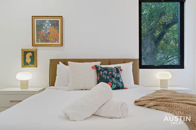 Bright modern vacation-rental bedroom with crisp white linens, rolled towels on the bed, textured cream and botanical accent pillows, tan headboard, matching bedside lamps, framed artwork, and a window view of leafy trees.