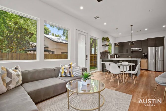 Sunlit Austin vacation rental open-plan living room and kitchen with gray sectional, geometric throw pillows, round glass coffee table, white dining chairs, and dark wood cabinets