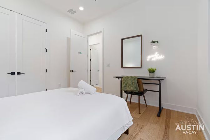 Bright minimalist Austin rental bedroom — crisp white bed with rolled towels, hardwood floors, compact desk and chair beneath a mirror, neon "good vibes" sign and small plants.