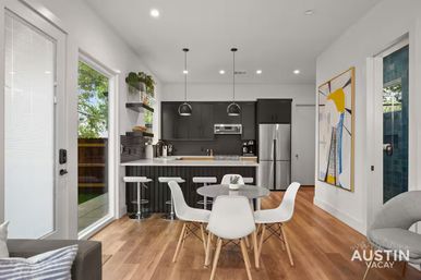 Bright modern Austin open-plan kitchen and dining area with hardwood floors, black cabinets, stainless refrigerator, breakfast bar with white stools, round table with white chairs, pendant lights, large glass door to patio and colorful wall art.