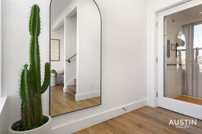 Tall potted cactus next to an arched full-length mirror in a bright modern entryway with white walls, light wood floors, a reflected staircase, and a glass door.