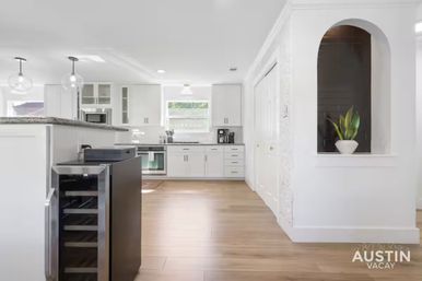 Bright modern white kitchen with island and granite countertop, globe pendant lights, open wine fridge, stainless appliances, wood-look floors, and arched niche with potted plant — airy Austin vacation rental kitchen.