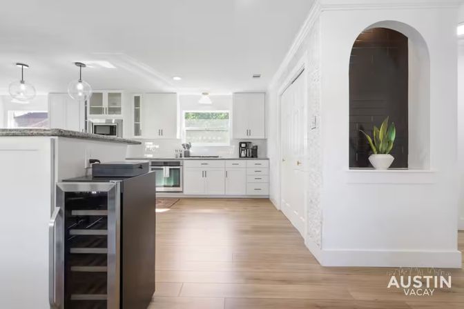 Bright modern white kitchen with island and granite countertop, globe pendant lights, open wine fridge, stainless appliances, wood-look floors, and arched niche with potted plant — airy Austin vacation rental kitchen.