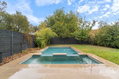 Rectangular backyard pool with attached spa, tan stone deck, dark wood privacy fence and leafy trees under a blue Austin sky