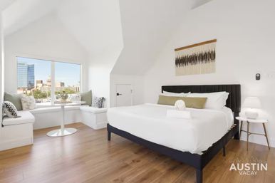 Bright modern vacation rental bedroom with king bed, cozy window seat and small table, white walls, hardwood floors and a large window framing a downtown city skyline view.