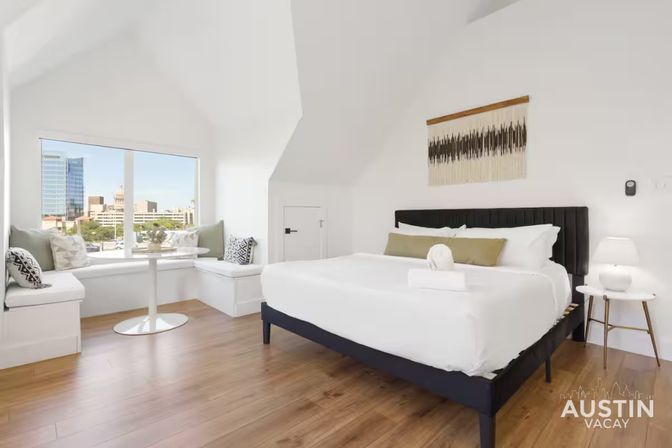 Bright modern vacation rental bedroom with king bed, cozy window seat and small table, white walls, hardwood floors and a large window framing a downtown city skyline view.
