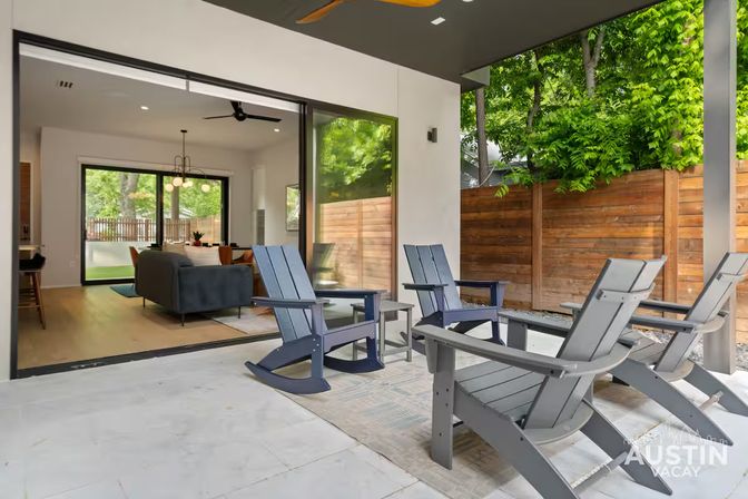 Covered modern backyard patio with four gray Adirondack rocking chairs and small side table, open sliding glass doors to a bright living and dining area, wooden privacy fence and lush green trees