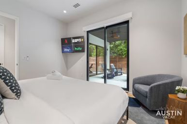 Bright modern bedroom with white bedding, wall-mounted TV displaying streaming app icons, sliding glass doors to a private patio with lounge chairs, gray armchair and wooden side table with plant.