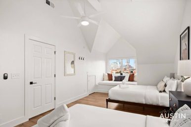 Bright airy attic bedroom with sloped ceiling, two white beds, cozy window seat with throw pillows overlooking a city skyline, hardwood floors and modern ceiling fan.