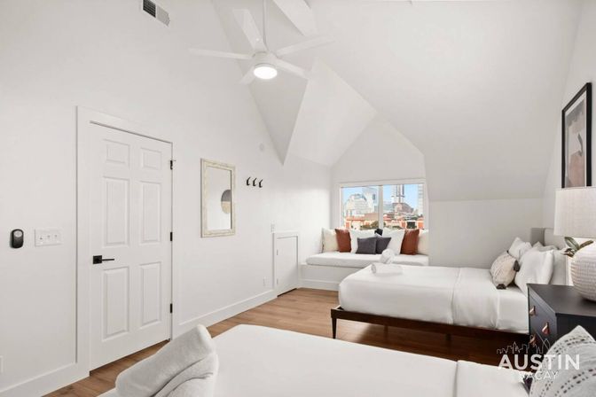 Bright airy attic bedroom with sloped ceiling, two white beds, cozy window seat with throw pillows overlooking a city skyline, hardwood floors and modern ceiling fan.