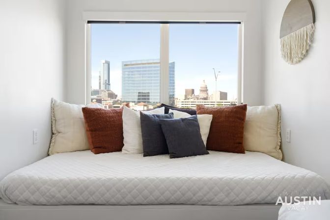 Cozy window daybed with quilted white bedding and rust, cream, and charcoal throw pillows, framed by a large window overlooking the Austin downtown skyline with a modern glass tower and capitol dome.