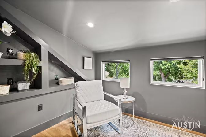 Cozy modern gray loft reading nook with white cushioned armchair, round side table and lamp, built-in shelves with plants and baskets, and two low windows framing green treetops.