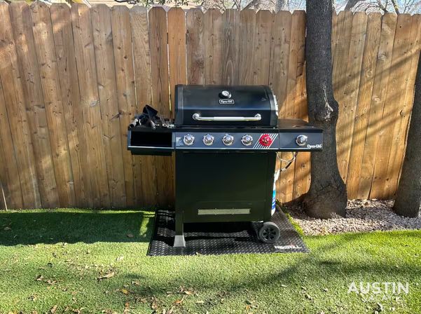Black five-burner gas grill on a diamond-plate mat over artificial turf, set against a wooden fence and tree in a sunny backyard