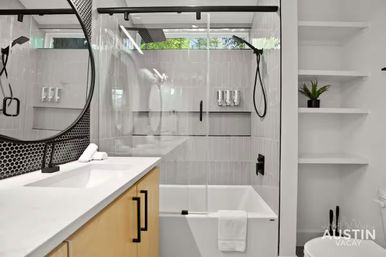 Modern Austin bathroom with glass-enclosed shower and rainfall head, white soaking tub with towel, light wood vanity, round mirror, black fixtures and open shelving