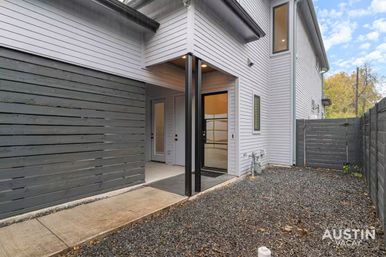 Contemporary two-story townhouse courtyard with light gray horizontal siding, covered entry supported by black metal posts, black-framed glass door, concrete walkway and gravel yard enclosed by dark horizontal slat privacy fence.